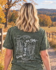 A woman viewed from behind walking on a gravel path in a rural outdoor setting, wearing a dark olive green acid-wash graphic t-shirt with cowboy boots and scripture printed on the back.