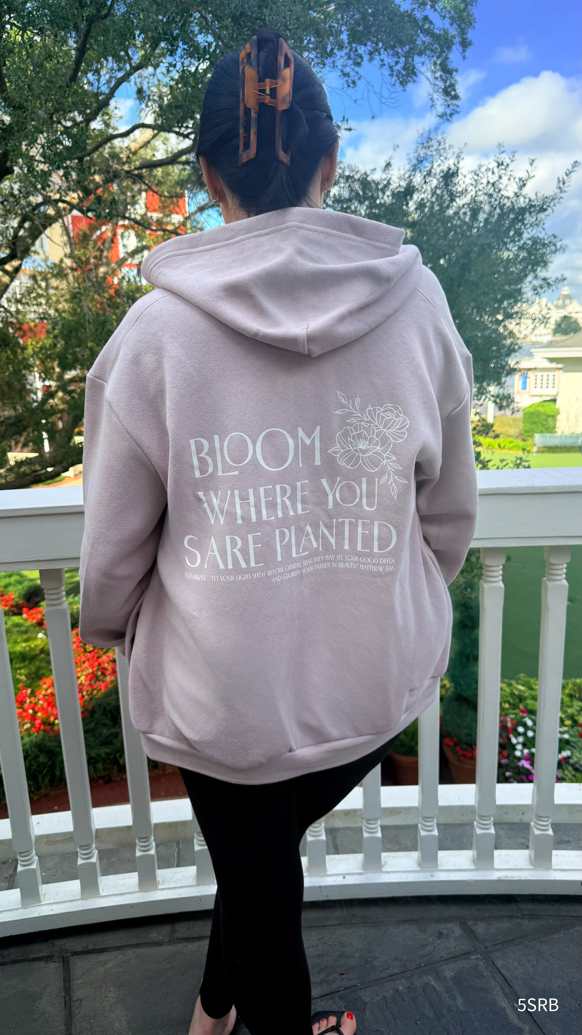 Person wearing a hoodie with 'Bloom Where You Are Planted' text on a balcony.