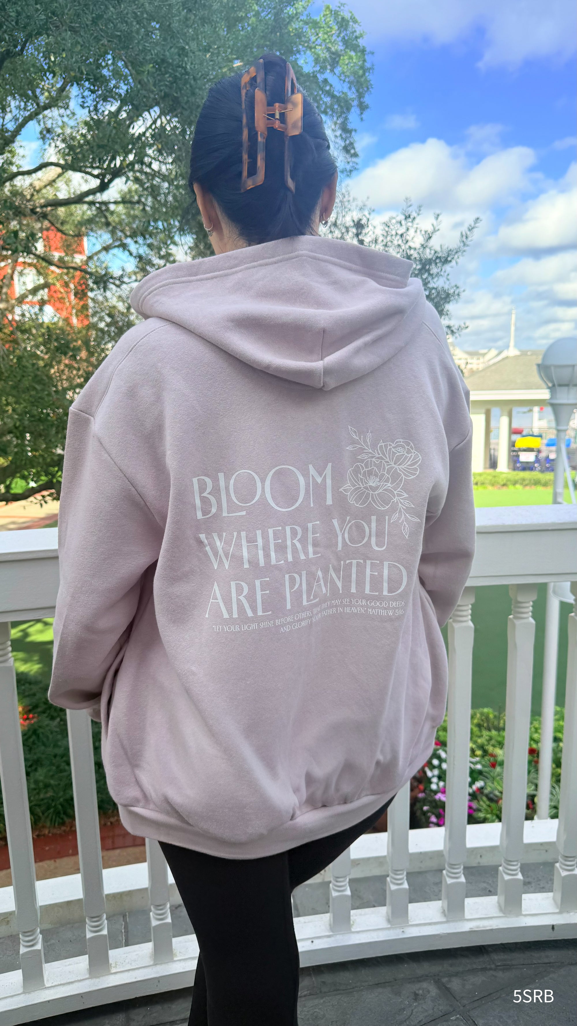 Person wearing a hoodie with 'Bloom Where You Are Planted' text on a balcony.