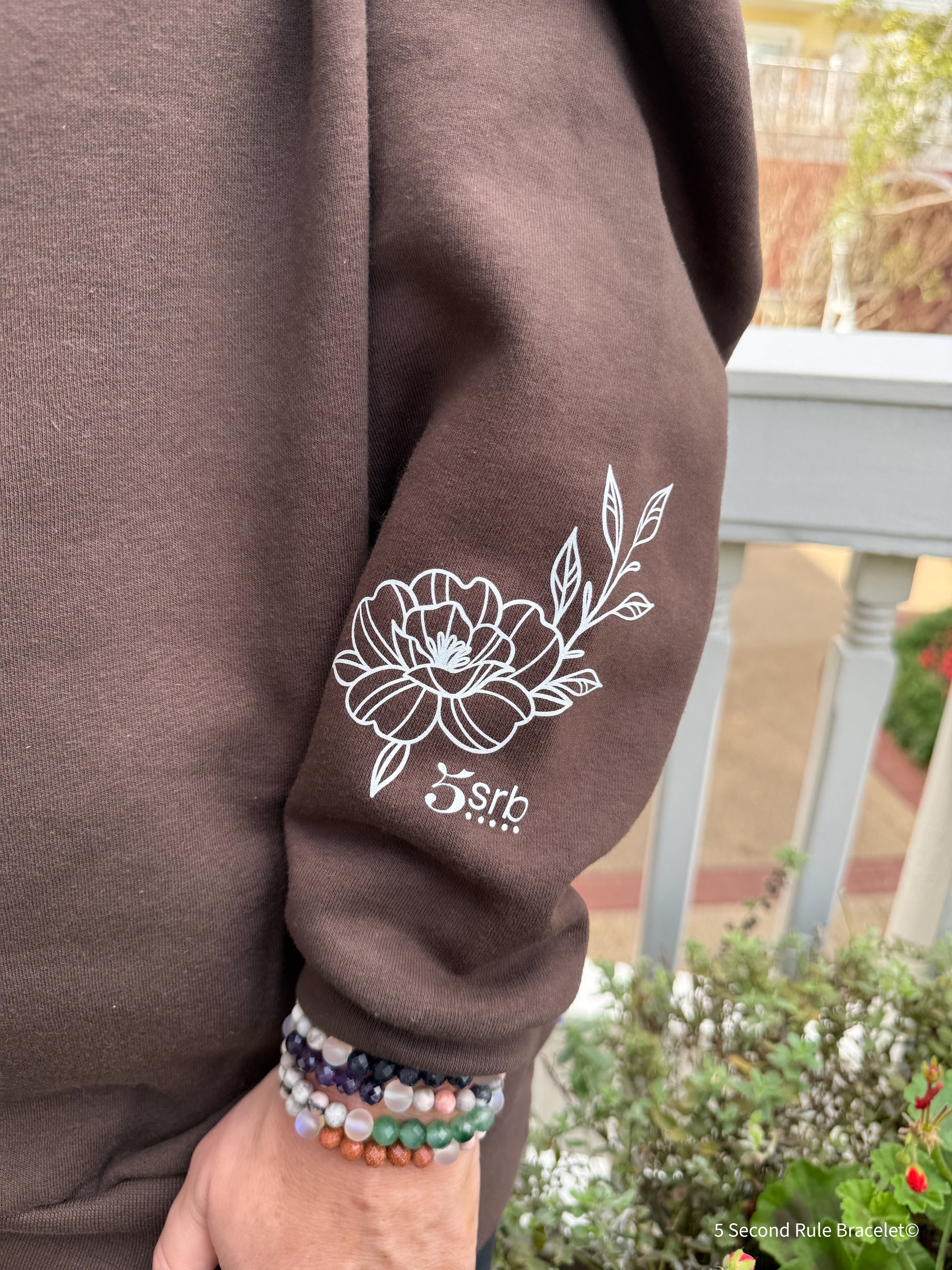 Brown sweatshirt with floral design on sleeve, held by a person wearing colorful bracelets.