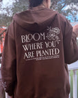 Person wearing a brown hoodie with 'Bloom Where You Are Planted' text on the back.