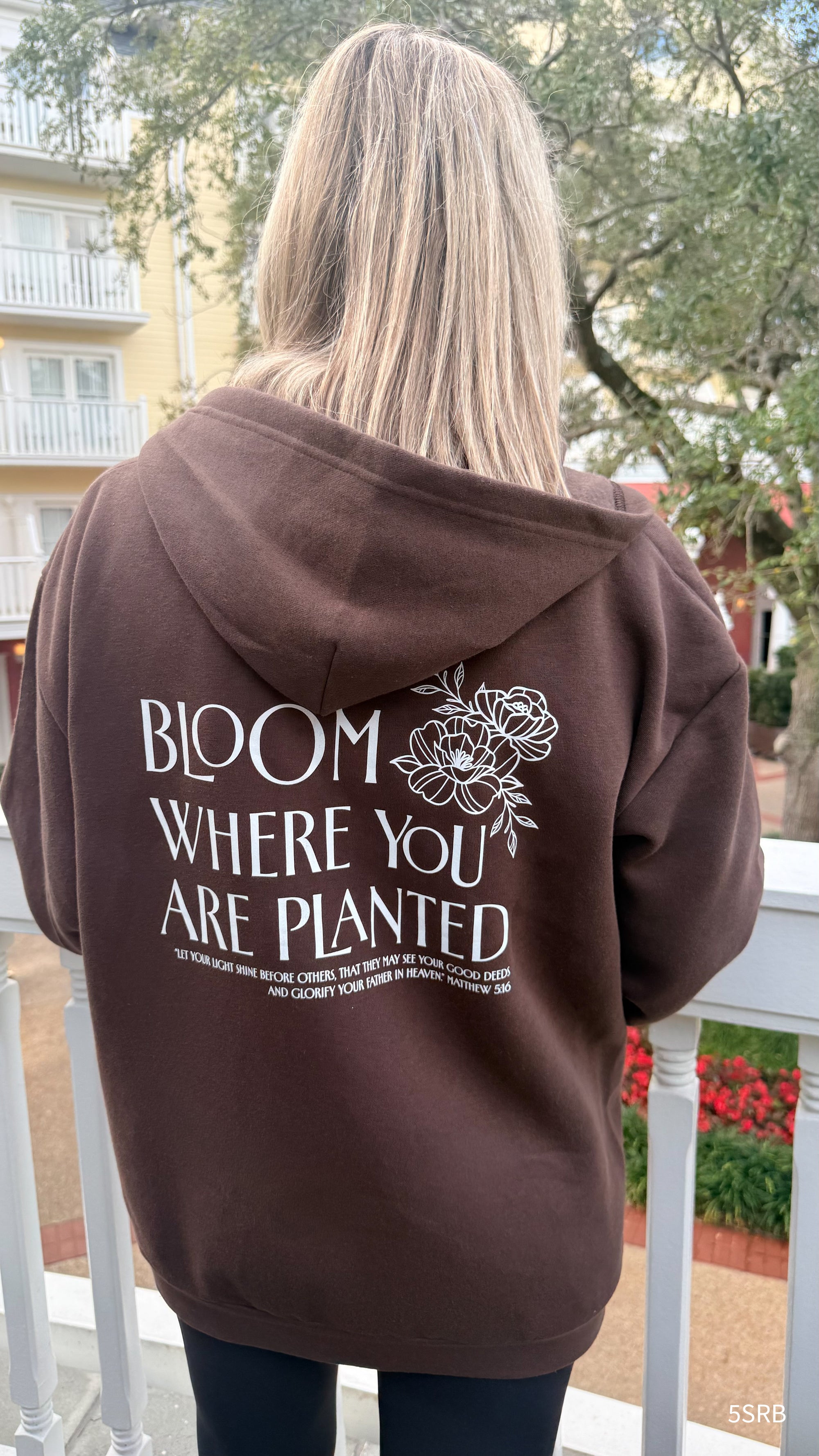 Person wearing a brown hoodie with 'Bloom Where You Are Planted' text and floral design on the back, standing outdoors.