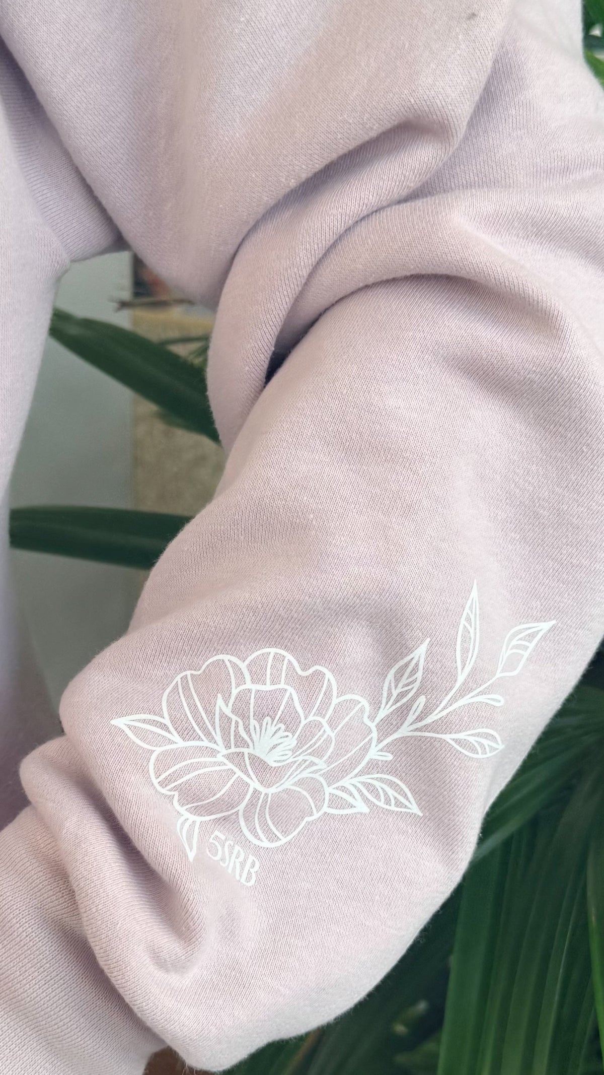 Light gray sweatshirt with white floral embroidery against a green plant background