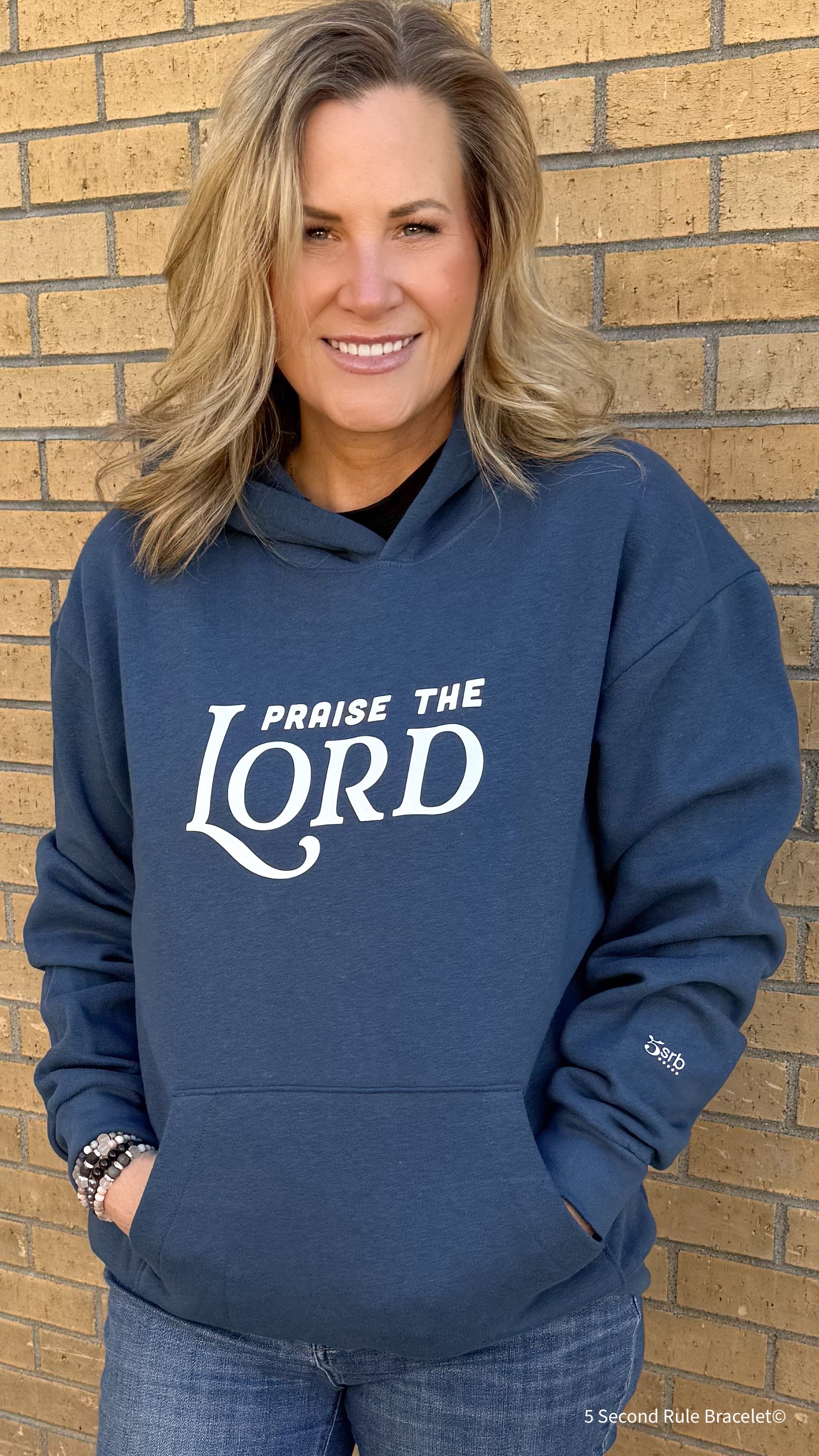 Woman wearing a blue hoodie with 'Praise the Lord' text against a brick wall.