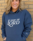 Woman wearing a blue hoodie with 'Praise the Lord' text against a brick wall.