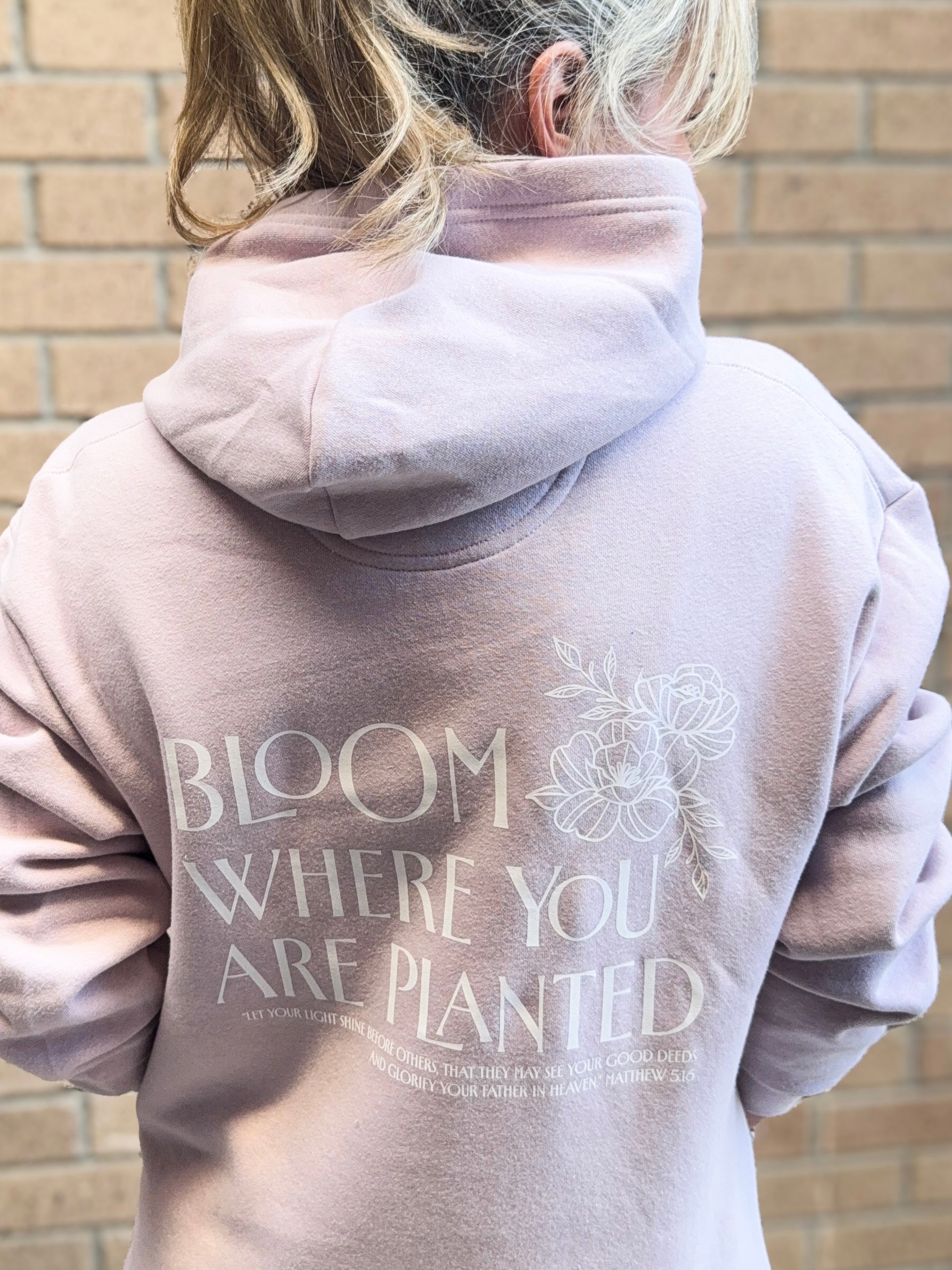 Person wearing a hoodie with 'Bloom Where You Are Planted' text on the back, standing against a brick wall.