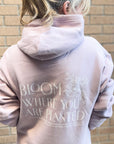 Person wearing a hoodie with 'Bloom Where You Are Planted' text on the back, standing against a brick wall.
