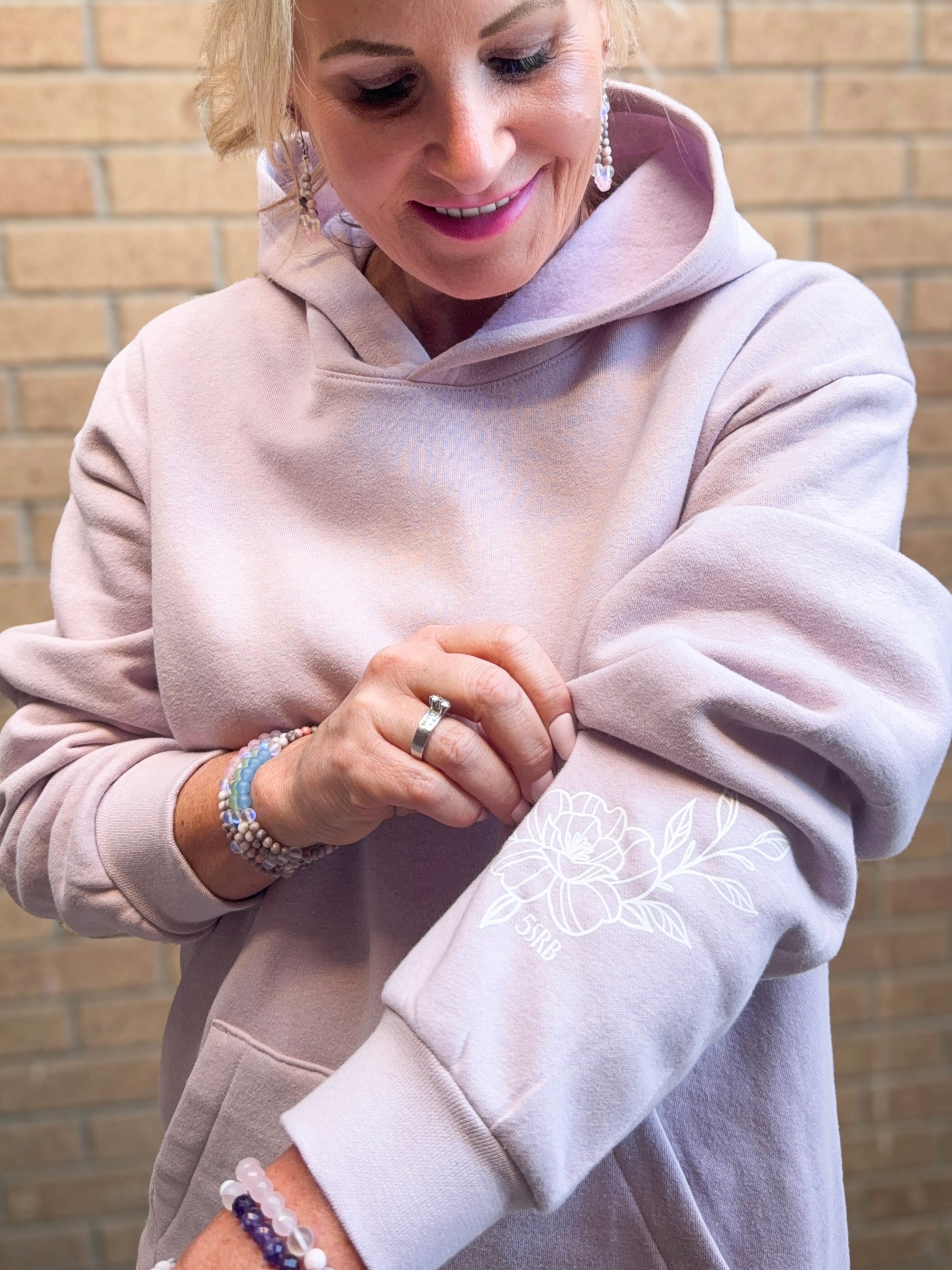 Woman wearing a light pink hoodie with floral embroidery, standing against a brick wall.