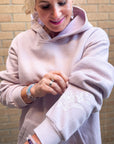 Woman wearing a light pink hoodie with floral embroidery, standing against a brick wall.