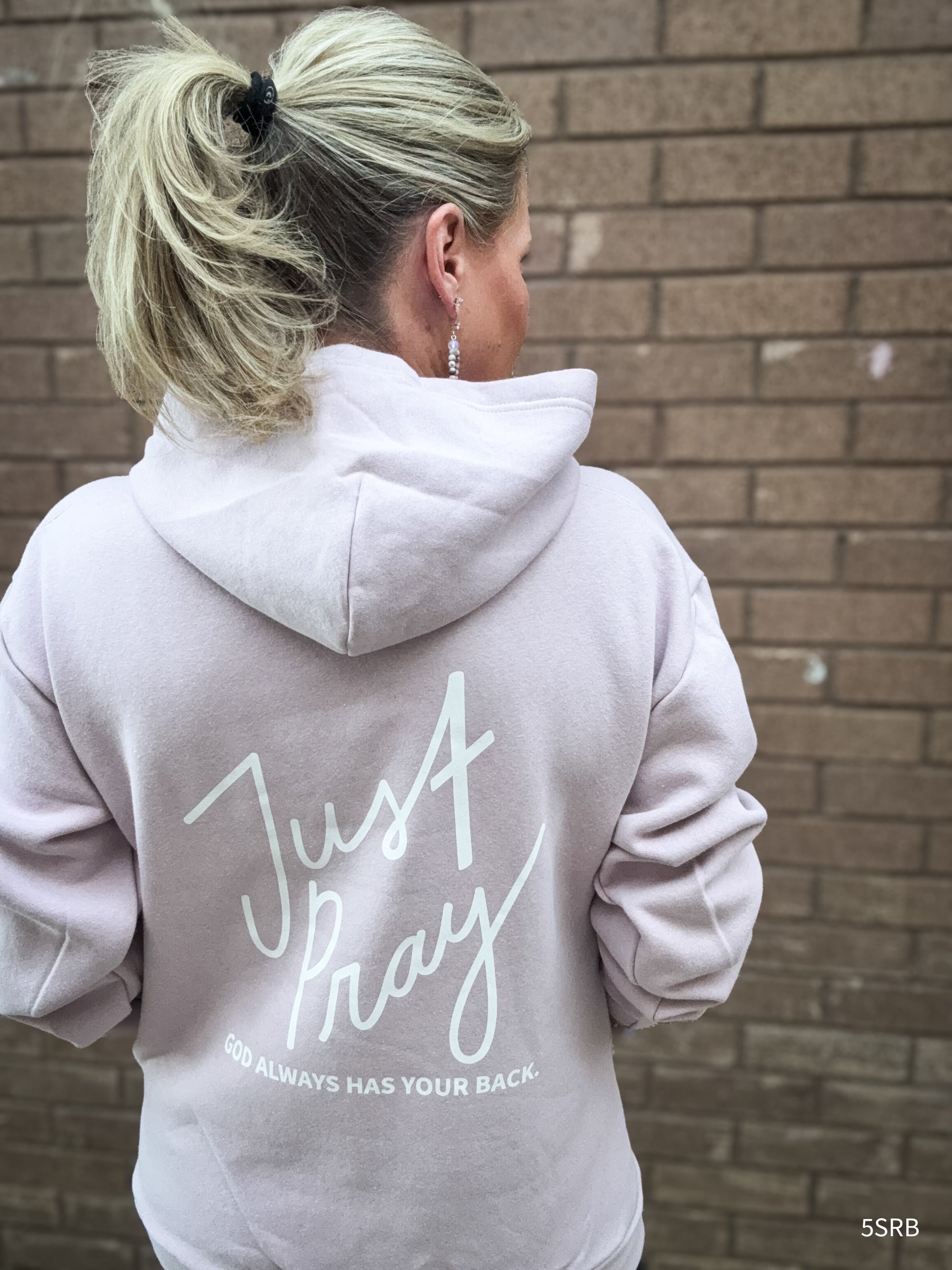 Person wearing a light gray hoodie with 'Just Pray' text on the back, standing against a brick wall.