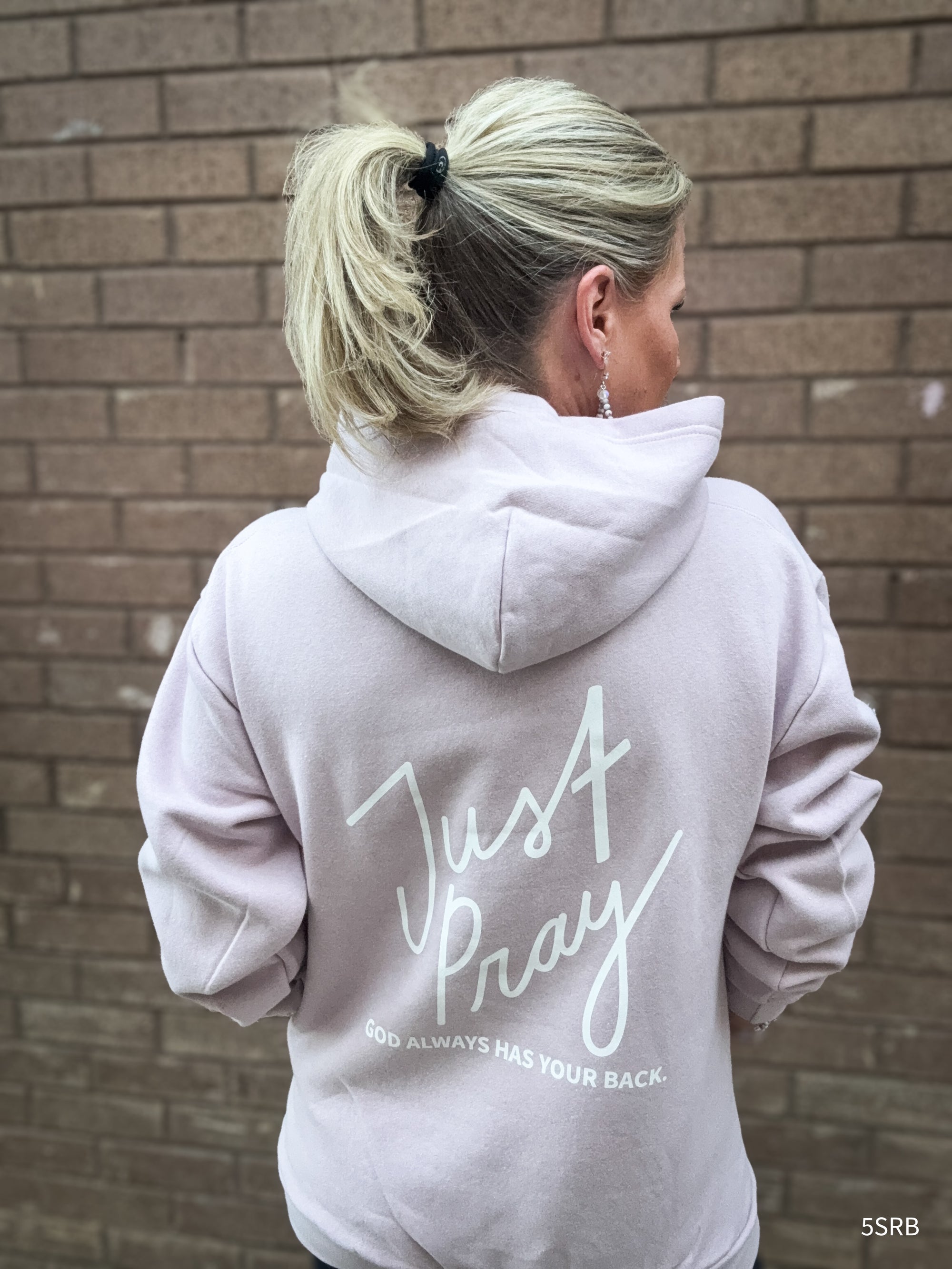 Person wearing a light gray hoodie with 'Just Pray' text on the back, standing against a brick wall.