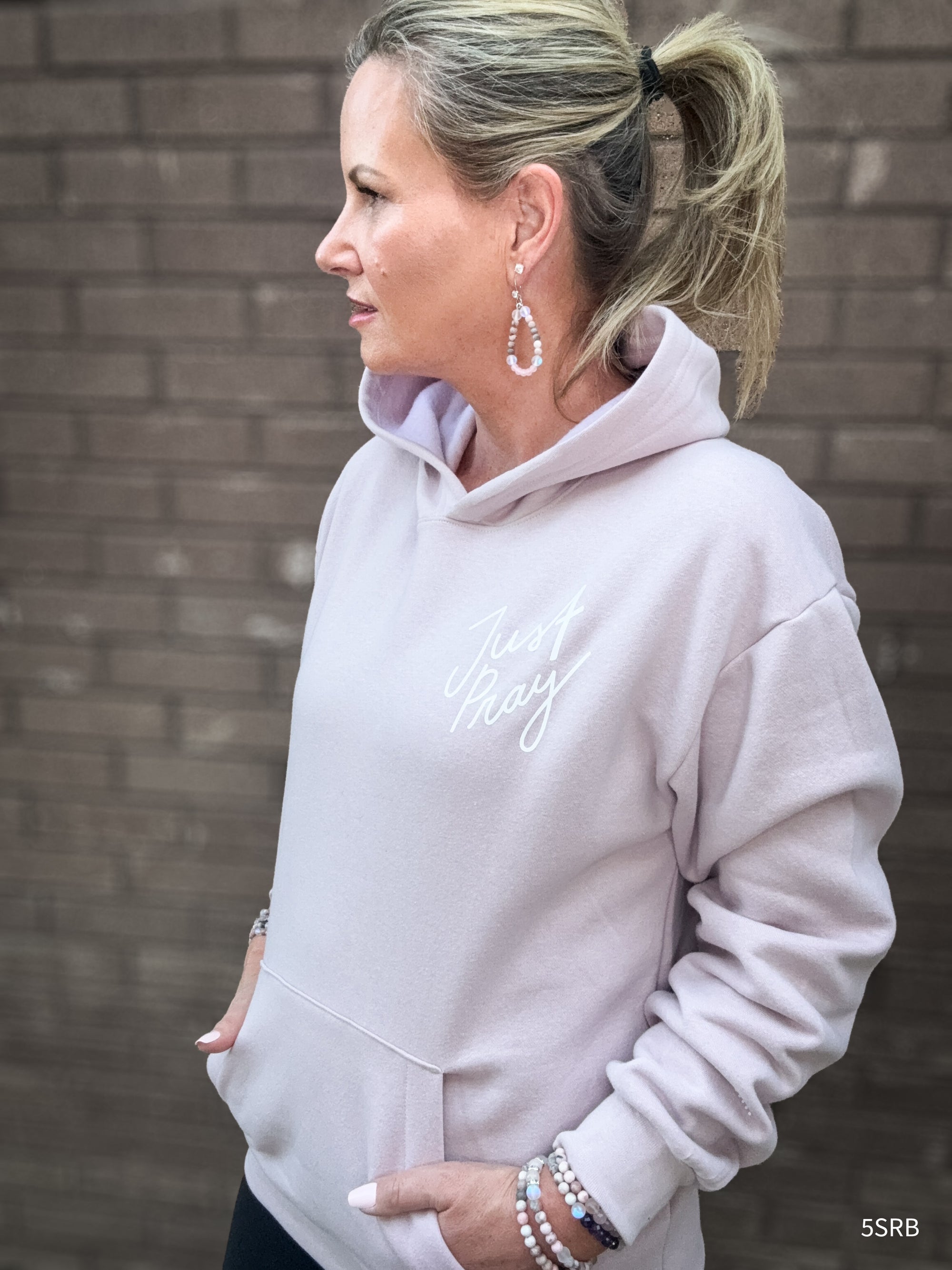 Woman wearing a light gray hoodie with text, standing against a brick wall.