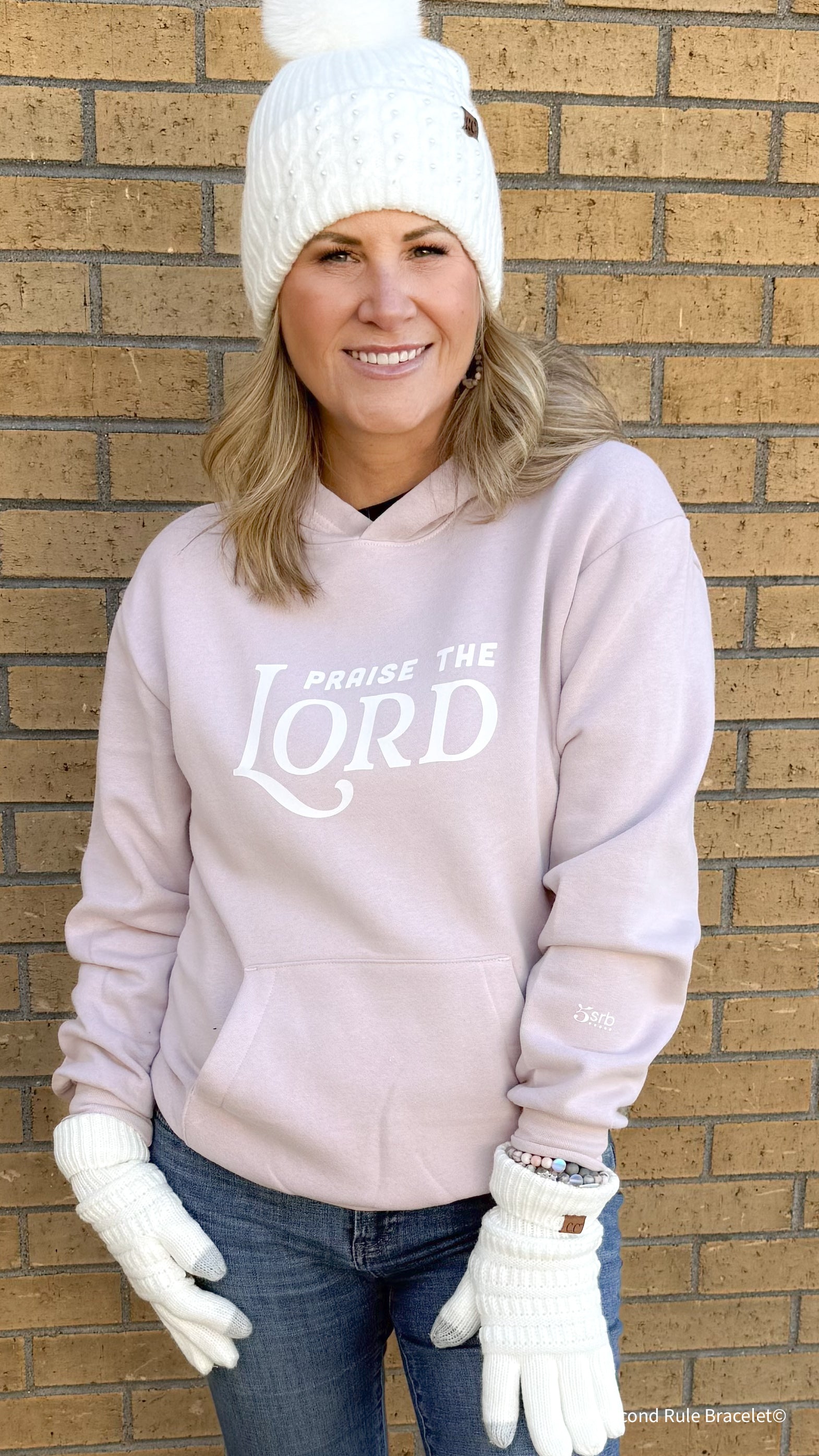 Person wearing a light pink hoodie with 'Praise the Lord' text, white beanie, and gloves against a brick wall.