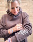 Woman wearing a brown hoodie with floral embroidery, standing against a brick wall.