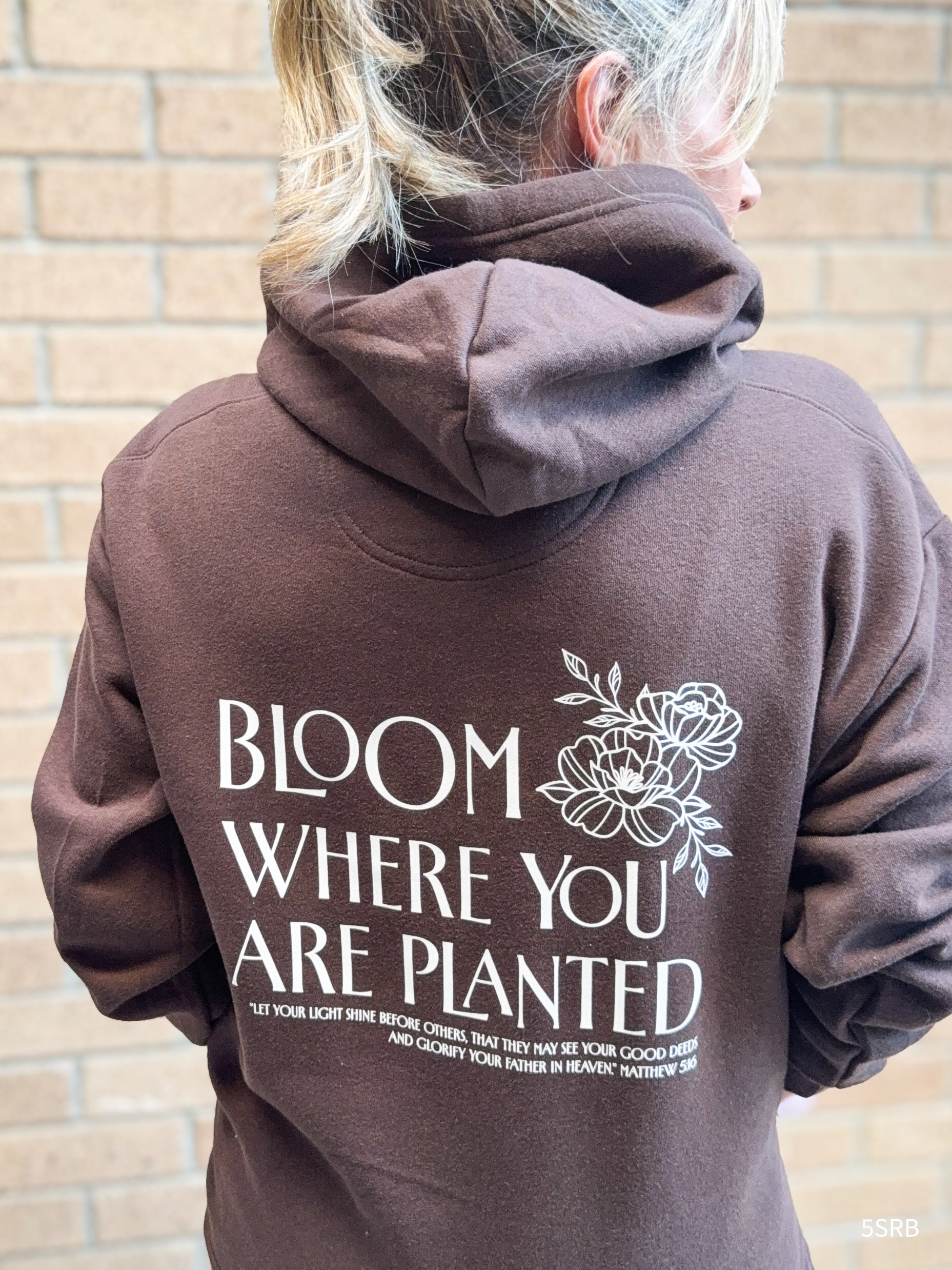 Person wearing a brown hoodie with 'Bloom Where You Are Planted' text and floral design on the back, standing against a brick wall.