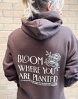 Person wearing a brown hoodie with 'Bloom Where You Are Planted' text and floral design on the back, standing against a brick wall.