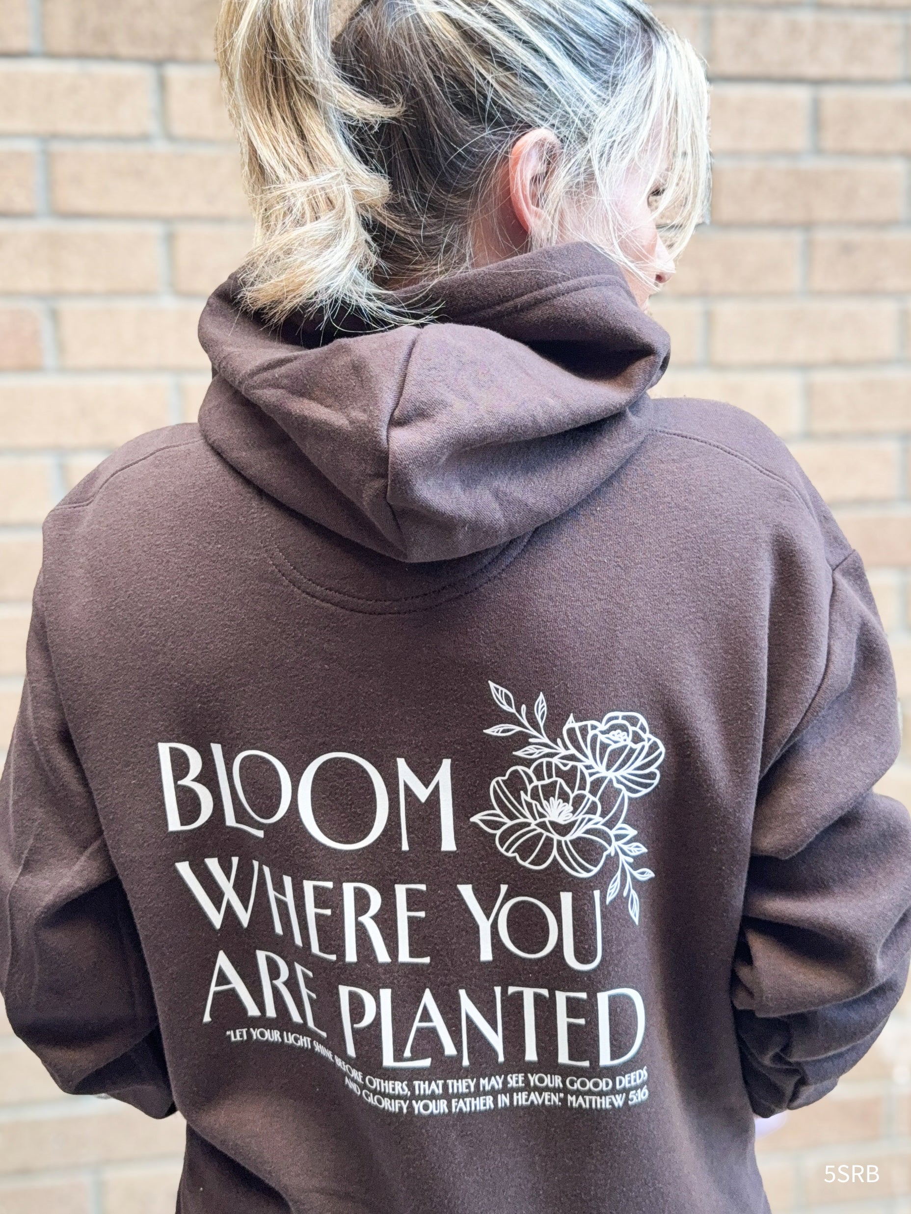 Person wearing a brown hoodie with 'Bloom Where You Are Planted' text and floral design on the back, standing against a brick wall.