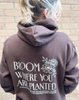 Person wearing a brown hoodie with 'Bloom Where You Are Planted' text and floral design on the back, standing against a brick wall.