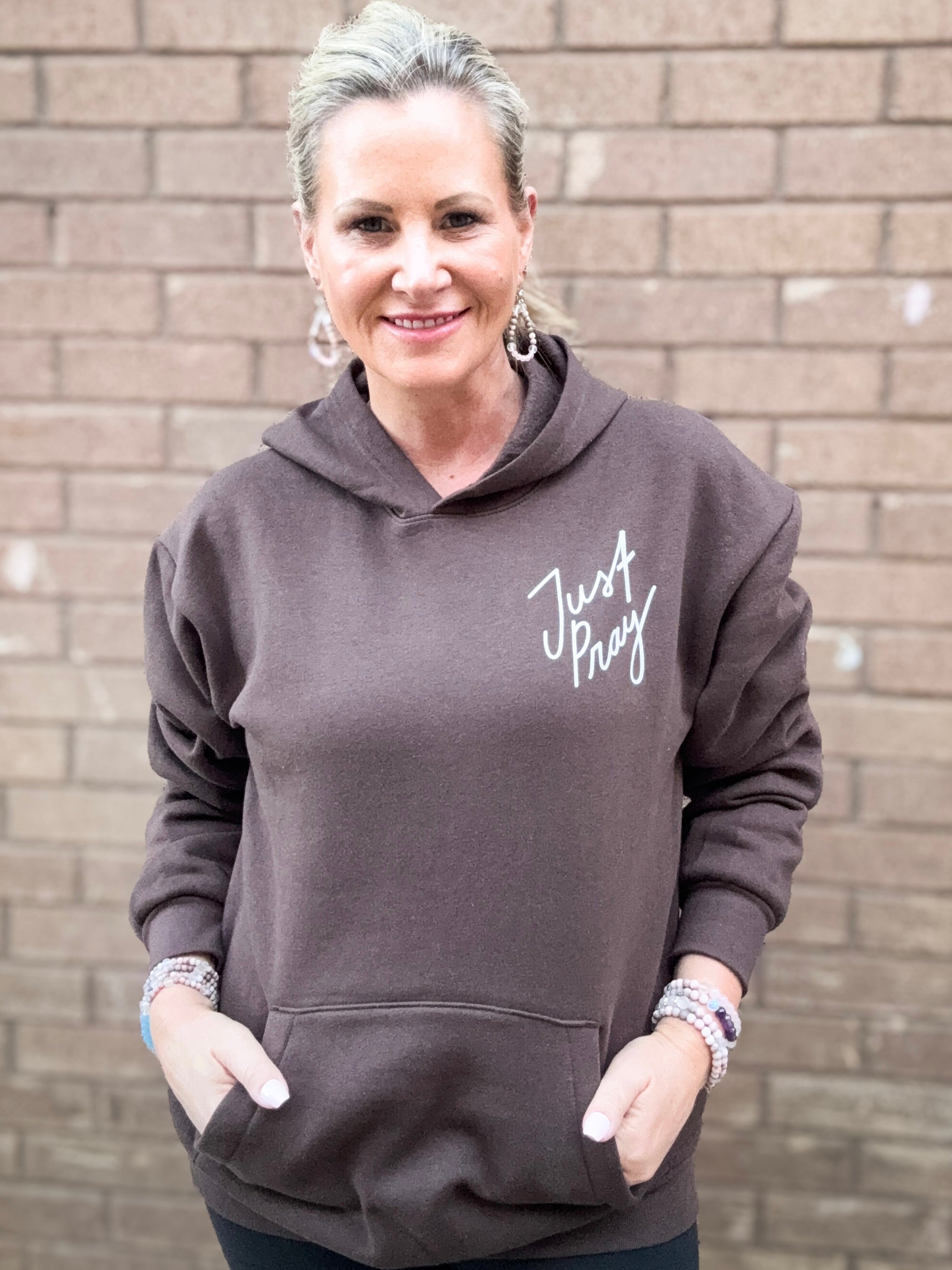 Woman wearing a brown hoodie with 'Just Pray' text against a brick wall.