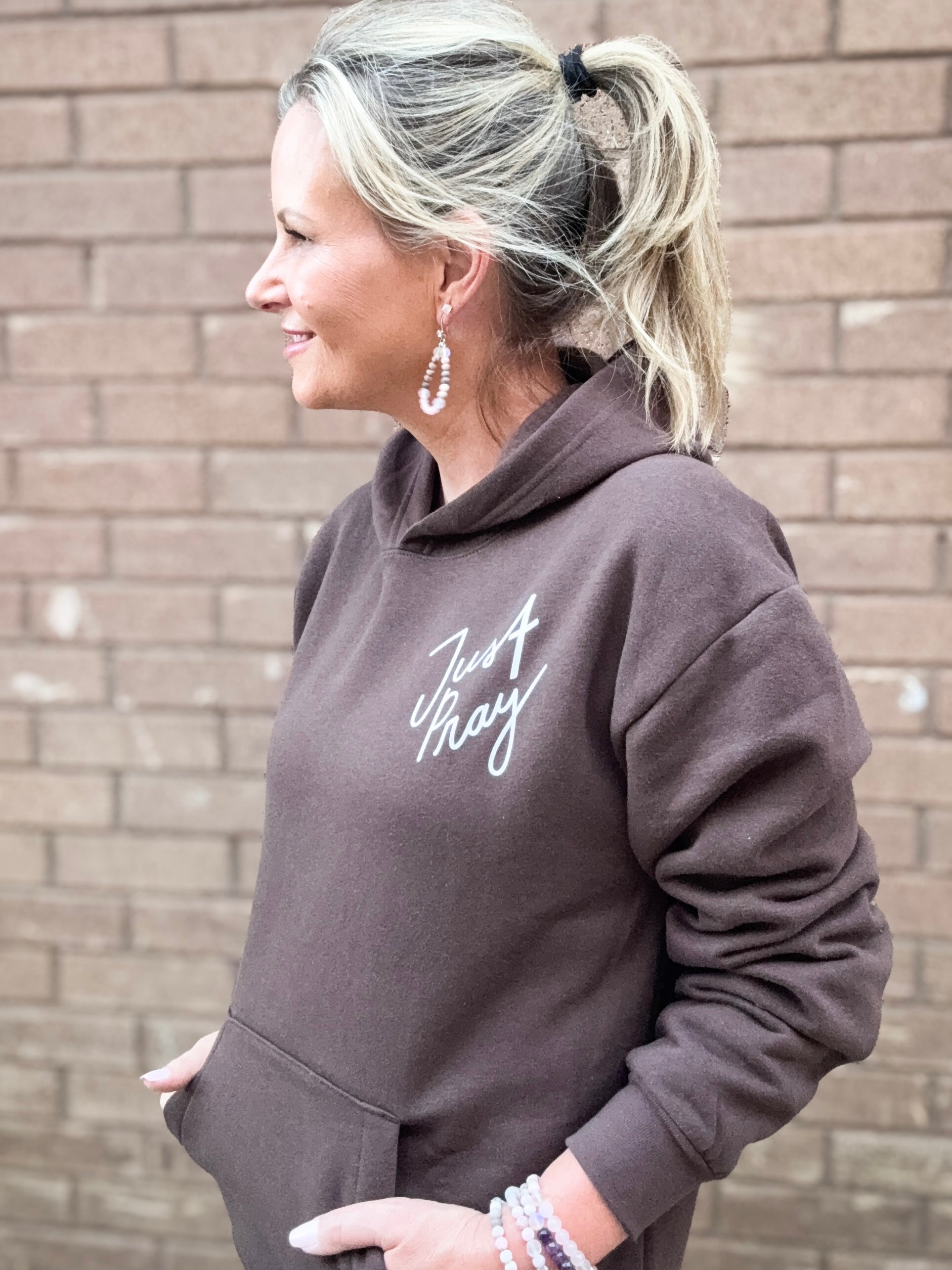 Woman wearing a brown hoodie with text, standing against a brick wall.