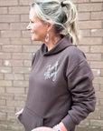 Woman wearing a brown hoodie with text, standing against a brick wall.