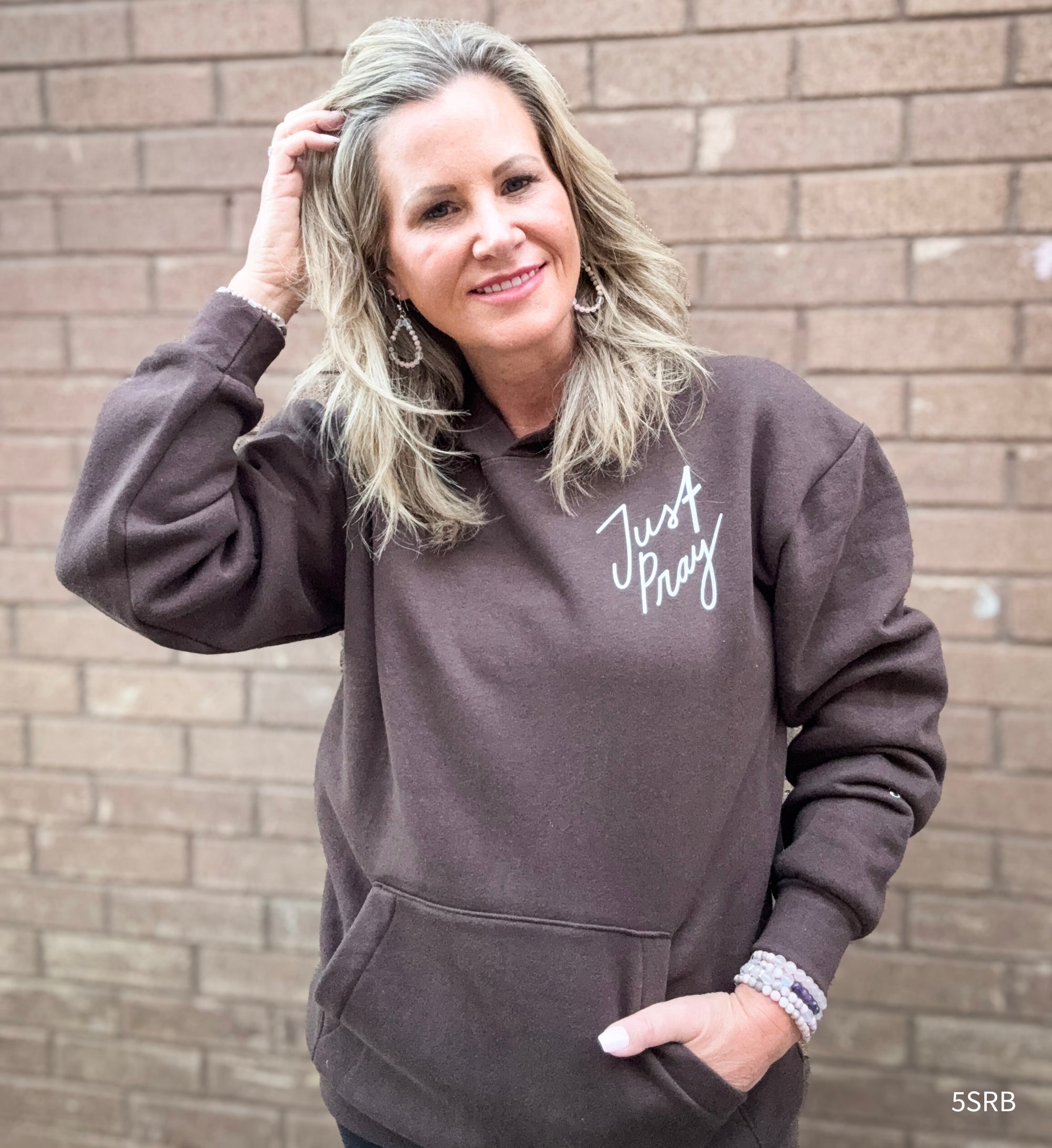 Woman wearing a brown hoodie with 'Just Pray' text against a brick wall.