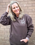 Woman wearing a brown hoodie with 'Just Pray' text against a brick wall.