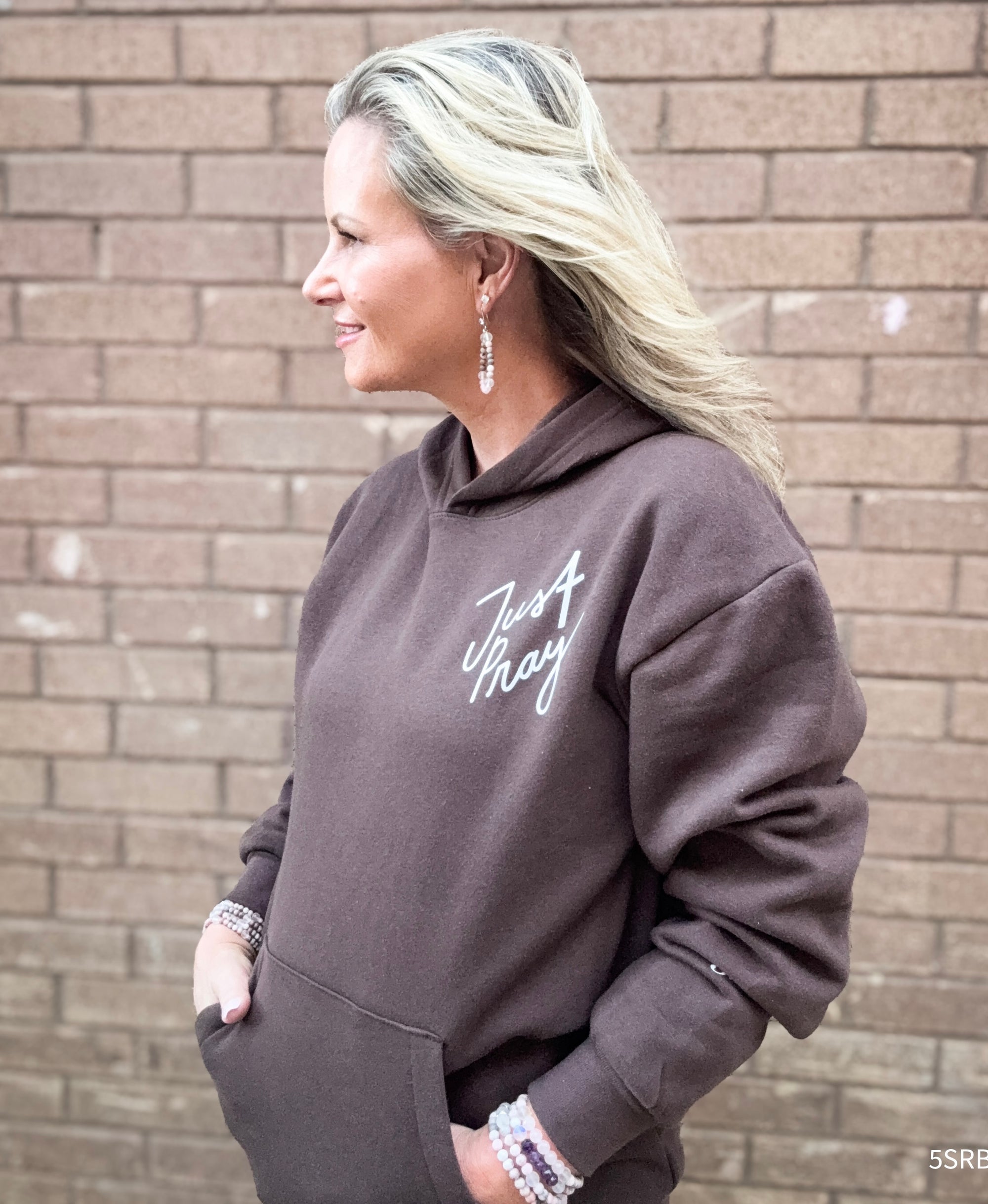Woman wearing a brown hoodie with text, standing against a brick wall.