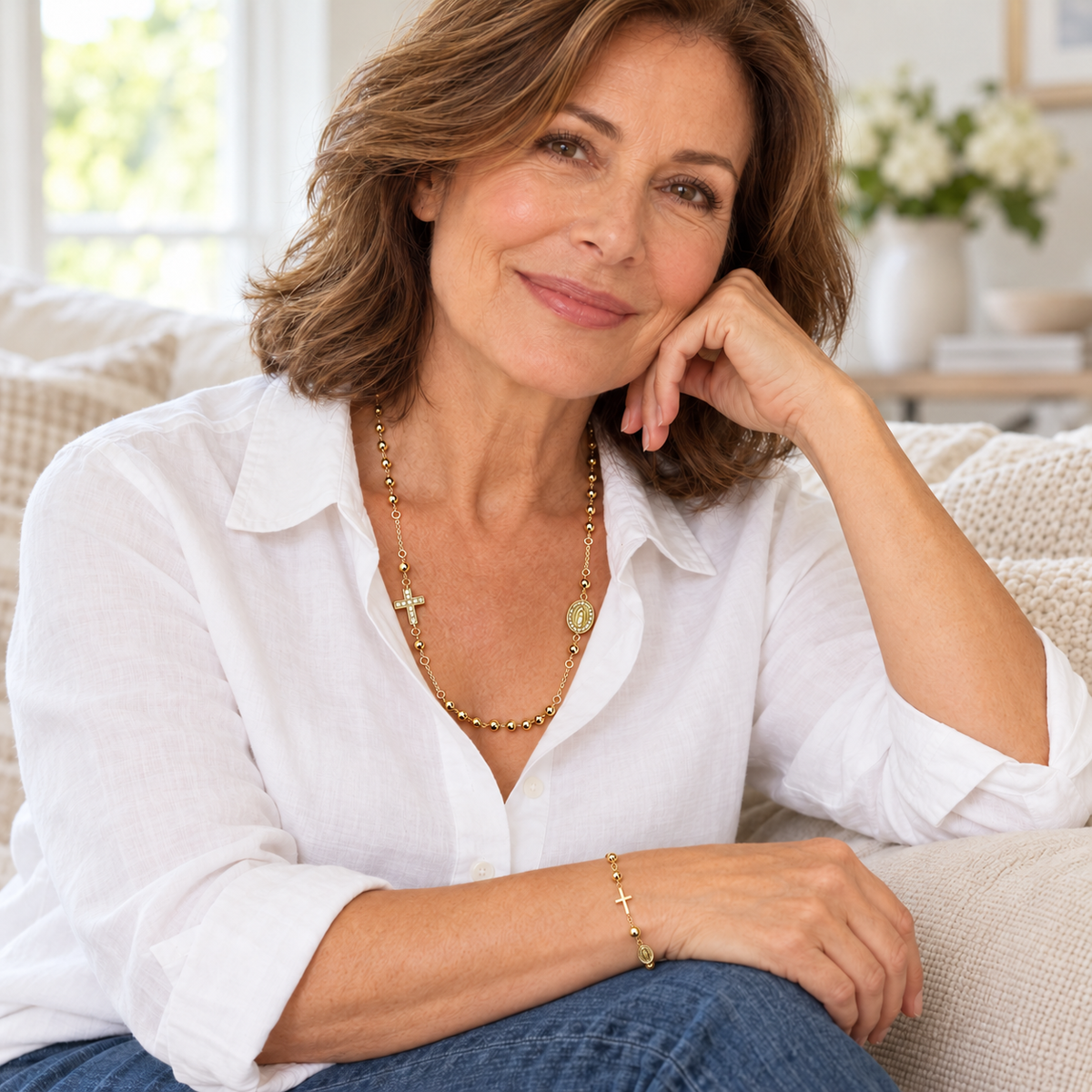 A woman with shoulder-length brown hair sits on a light beige couch, wearing a white button-up shirt, blue jeans, a gold beaded necklace with a cross pendant, and a matching gold bracelet.