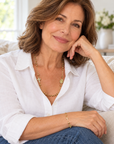 A woman with shoulder-length brown hair sits on a light beige couch, wearing a white button-up shirt, blue jeans, a gold beaded necklace with a cross pendant, and a matching gold bracelet.