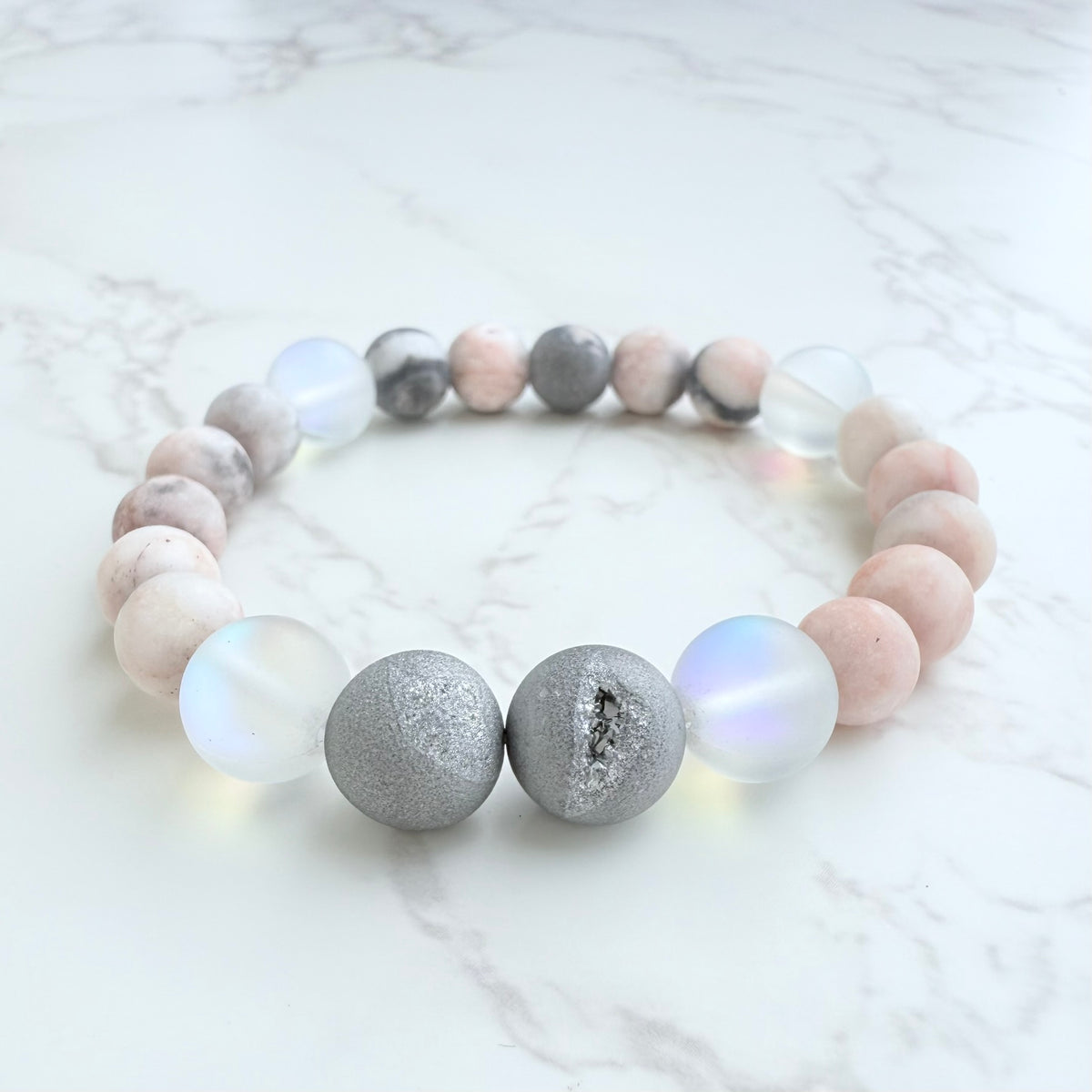 A bracelet with silver-colored druzies and pink zebra jasper beads on a marble surface.