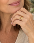 A woman with wavy brown hair wearing a cream knit sweater and a gold ring with a cross design, posing in a softly lit cafe setting.