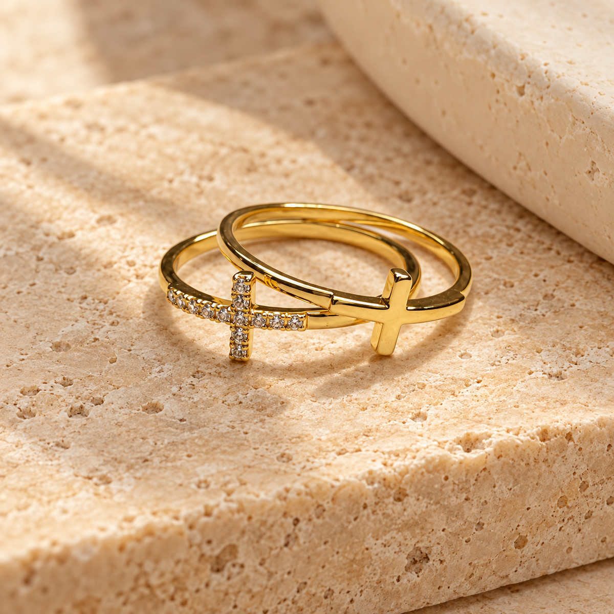 Two gold rings displayed on a textured beige stone surface. One ring features a polished cross with a row of small clear stones, and the other is a smooth open band.