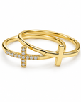 A pair of gold rings, one featuring a cross pavé-set with small clear stones and the other a solid cross pendant, displayed on a clean white background.