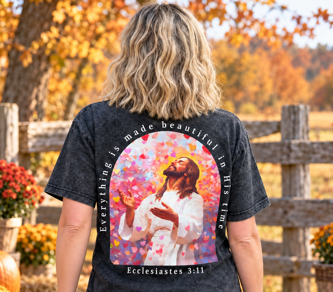 A woman stands outdoors in a fall yard wearing a black acid-wash graphic t-shirt. The back of the shirt features a large colorful print of Jesus surrounded by hearts, with the text 'Everything is made beautiful in His time' and 'Ecclesiastes 3:11'.