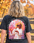 A woman stands outdoors in a fall yard wearing a black acid-wash graphic t-shirt. The back of the shirt features a large colorful print of Jesus surrounded by hearts, with the text 'Everything is made beautiful in His time' and 'Ecclesiastes 3:11'.