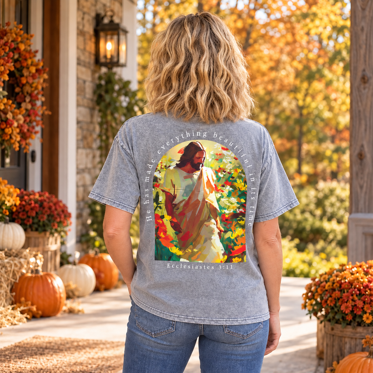 A woman stands on a porch wearing a heather gray graphic T-shirt from the back, featuring a colorful print of Jesus in a garden with the text 'he has made everything beautiful in its time