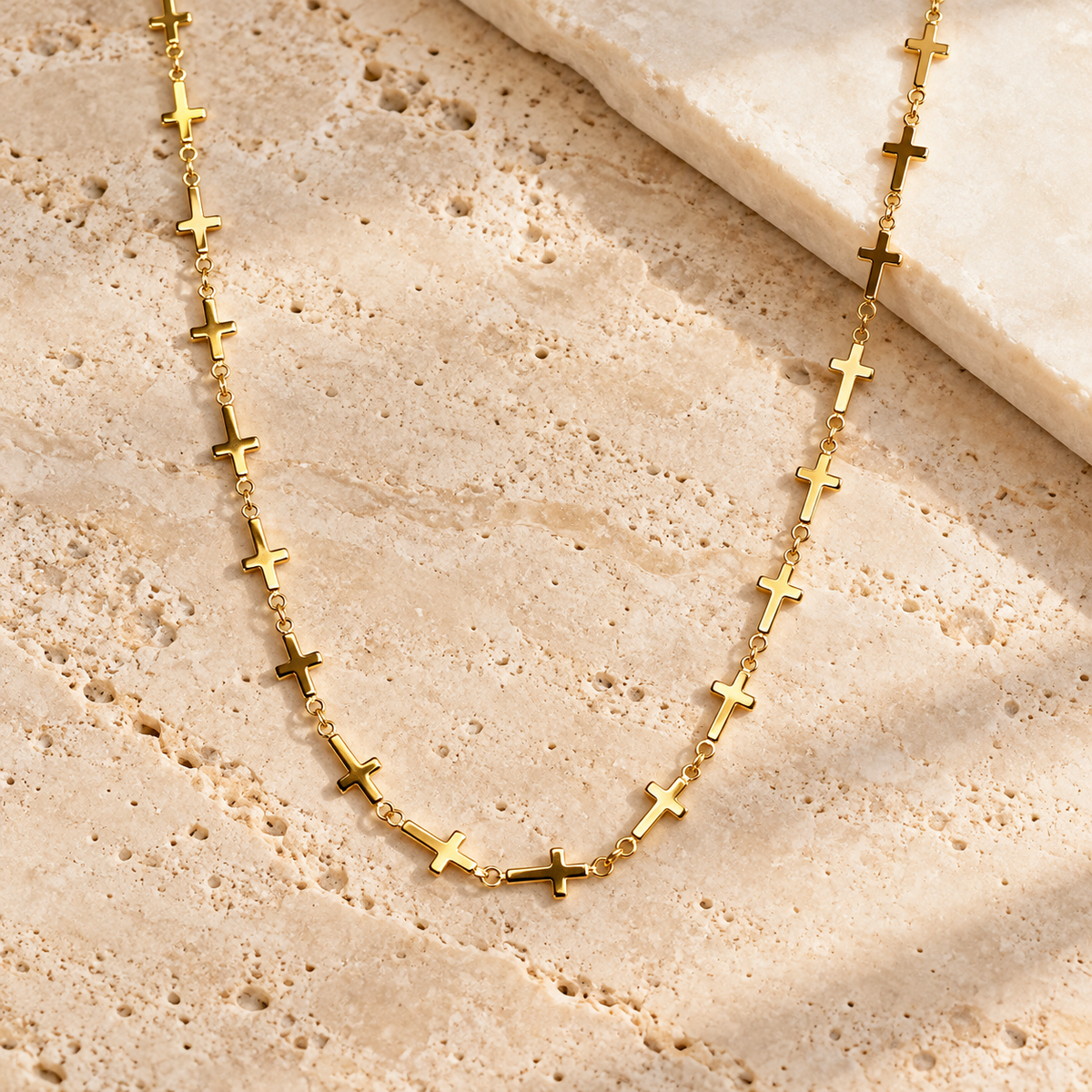 A gold chain necklace featuring multiple small cross charms spaced along the link chain, displayed on a textured beige stone surface.