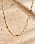A gold chain necklace featuring multiple small cross charms spaced along the link chain, displayed on a textured beige stone surface.