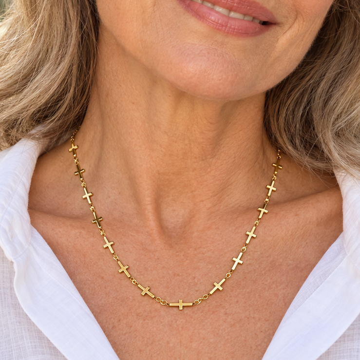 A smiling woman with blonde hair wearing a delicate gold cross necklace and a white collared shirt, photographed outdoors with a softly blurred beige stone background.