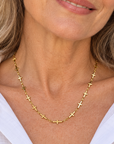 A smiling woman with blonde hair wearing a delicate gold cross necklace and a white collared shirt, photographed outdoors with a softly blurred beige stone background.