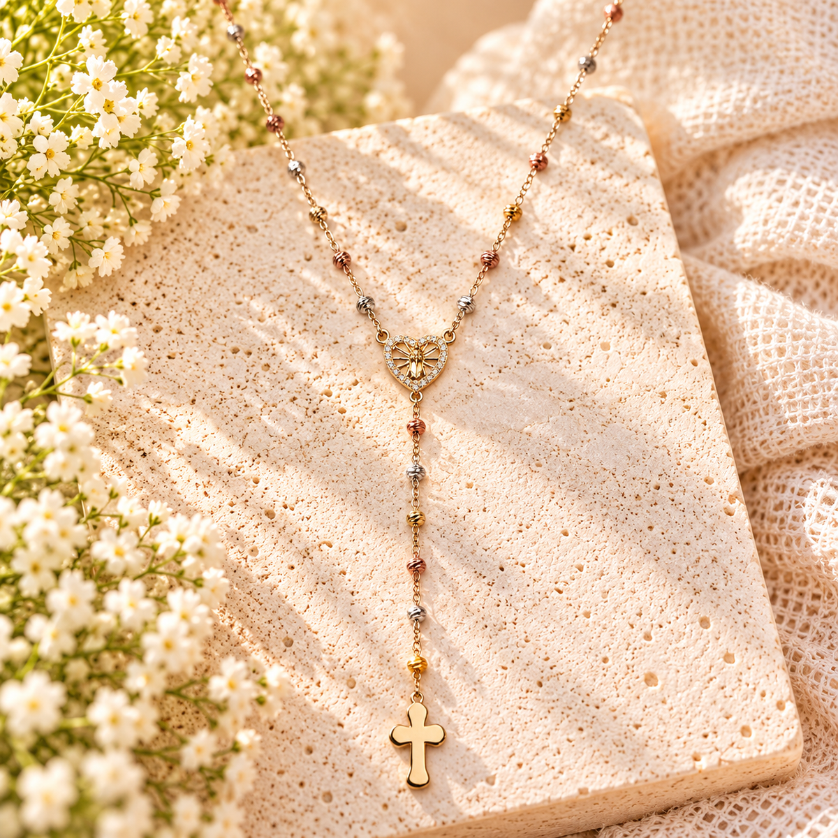 A delicate gold Y-necklace with a heart-shaped medallion and cross pendant, adorned with small multicolored beads, displayed on a textured beige stone slab with white flowers and cream fabric.