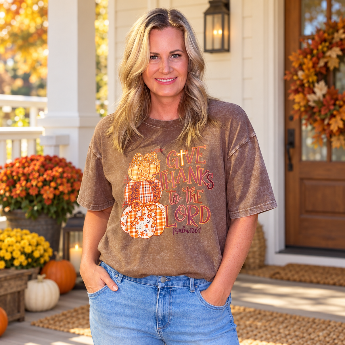 A woman stands on a front porch wearing a brown acid-wash graphic t-shirt with a fall-themed design that reads 'GIVE THANKS TO THE LORD' with plaid and orange pumpkin graphics. She is wearing blue jeans, with pumpkins, flowers, and a wooden door in the background.