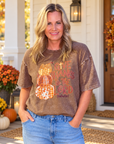 A woman stands on a front porch wearing a brown acid-wash graphic t-shirt with a fall-themed design that reads 'GIVE THANKS TO THE LORD' with plaid and orange pumpkin graphics. She is wearing blue jeans, with pumpkins, flowers, and a wooden door in the background.
