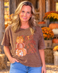 A woman wearing a brown acid-wash graphic t-shirt with a fall harvest design, standing in front of a rustic barn backdrop with pumpkins and mums.