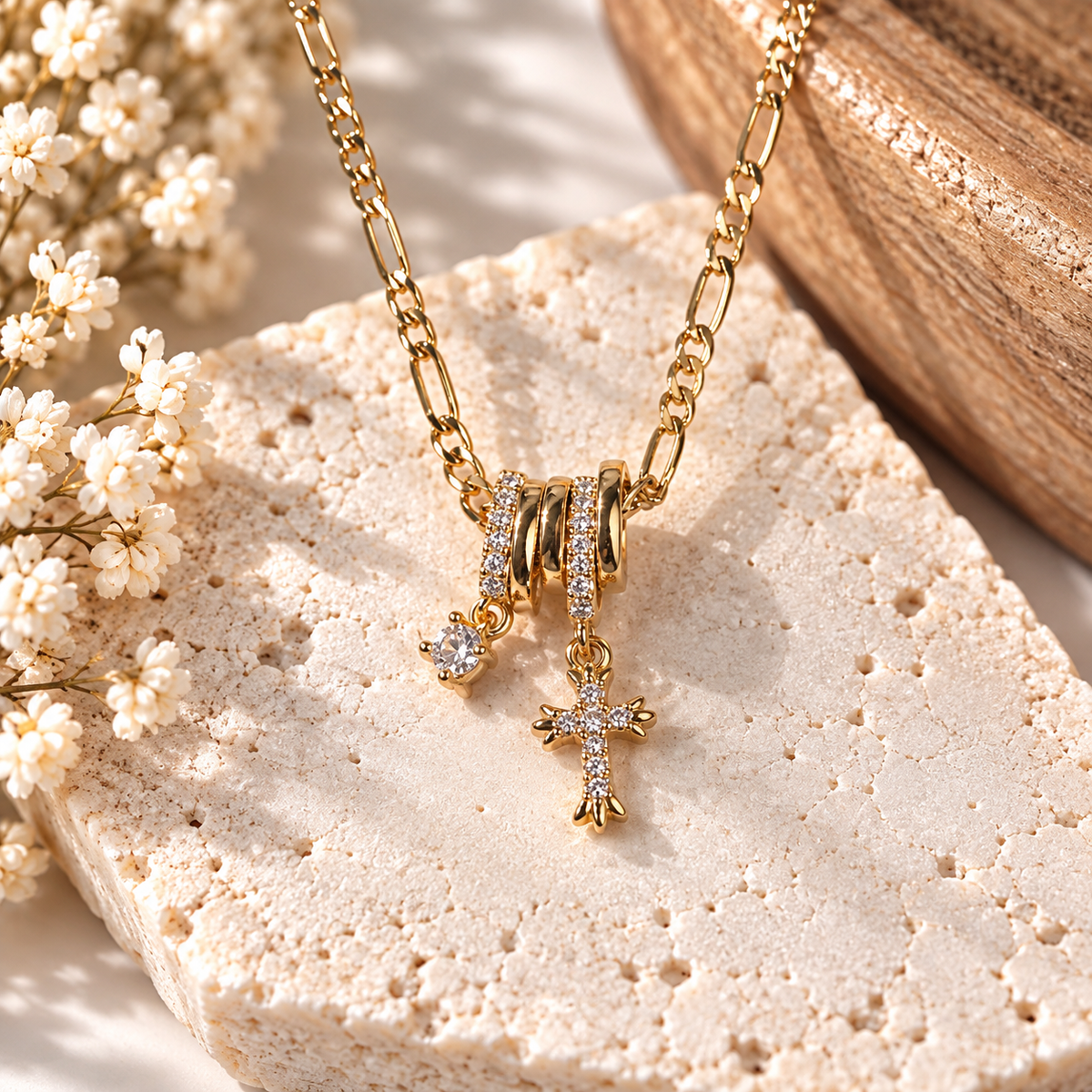 Gold cross pendant necklace with a small heart charm and a ring-shaped pendant, displayed on a textured stone slab with white flowers and a wooden prop.