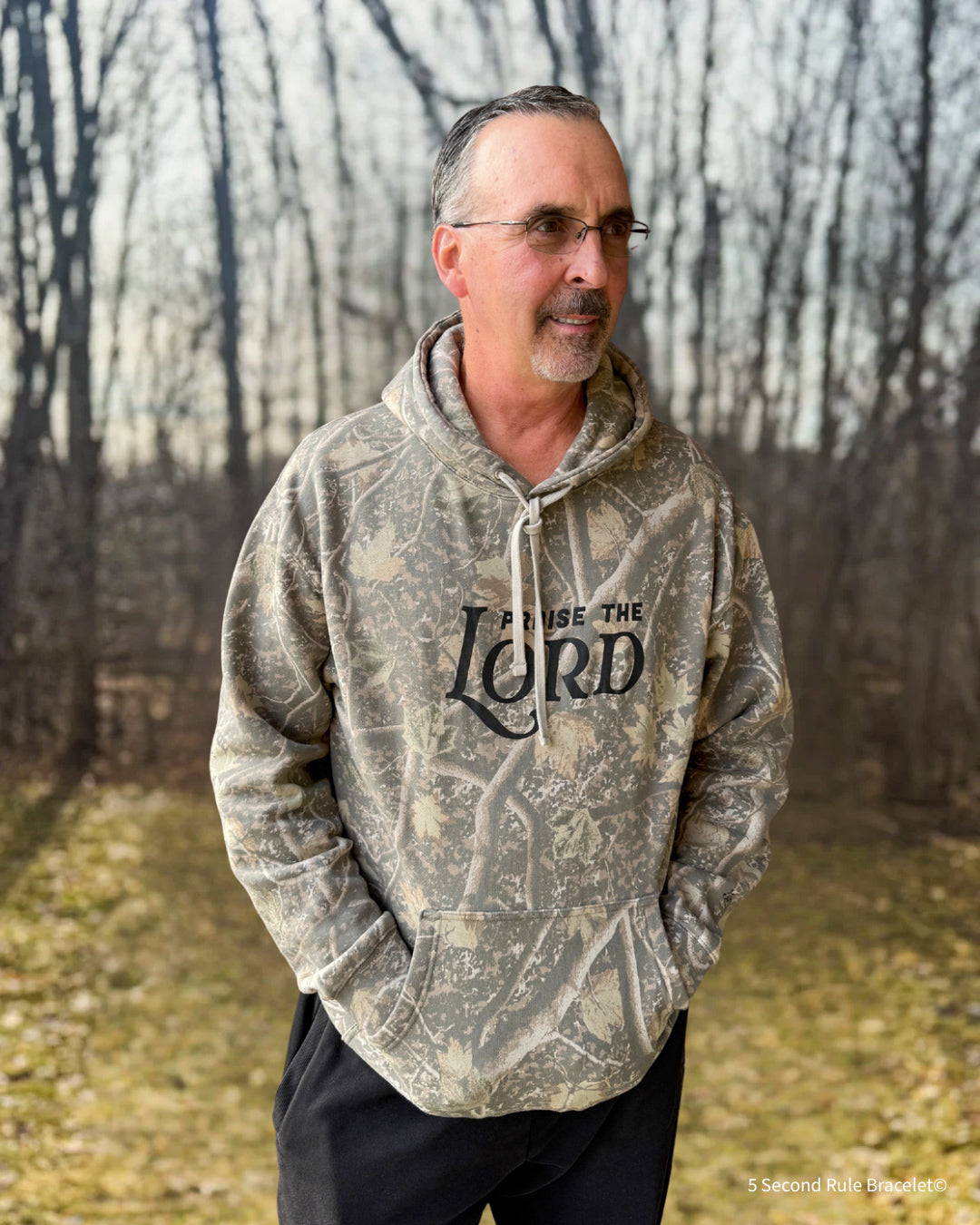 Man wearing a camouflage hoodie with text in a forest setting