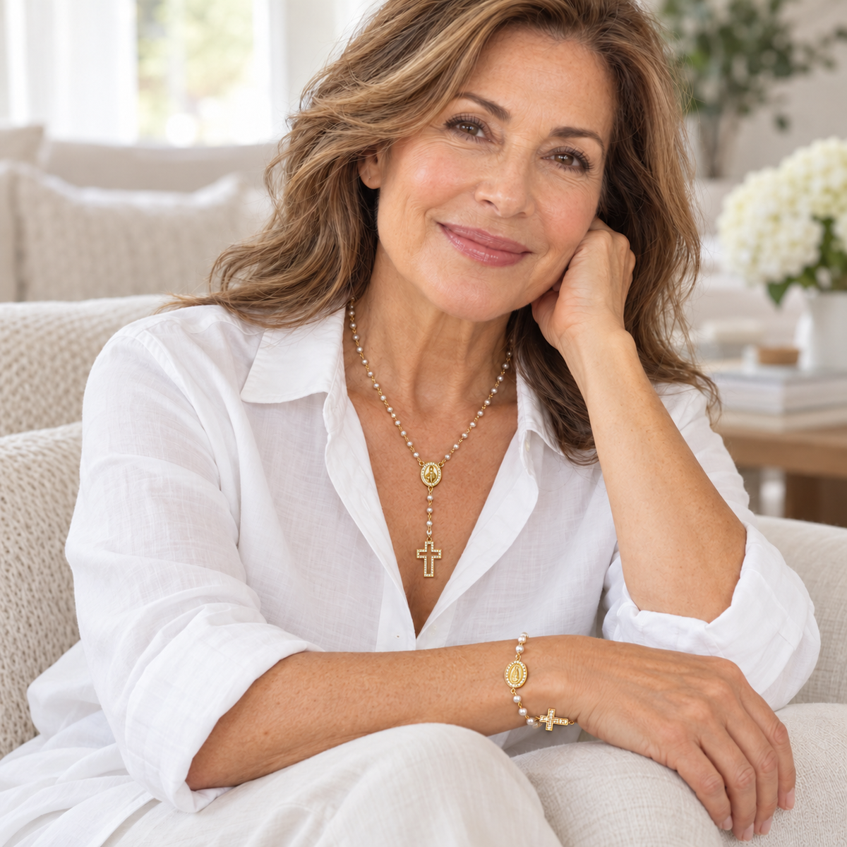 A woman with wavy brown hair sits on a light beige sofa wearing a white button-up shirt and white pants, styled with gold jewelry including a cross pendant necklace, a cross bracelet, and a ring.
