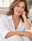 A woman with wavy brown hair sits on a light beige sofa wearing a white button-up shirt and white pants, styled with gold jewelry including a cross pendant necklace, a cross bracelet, and a ring.
