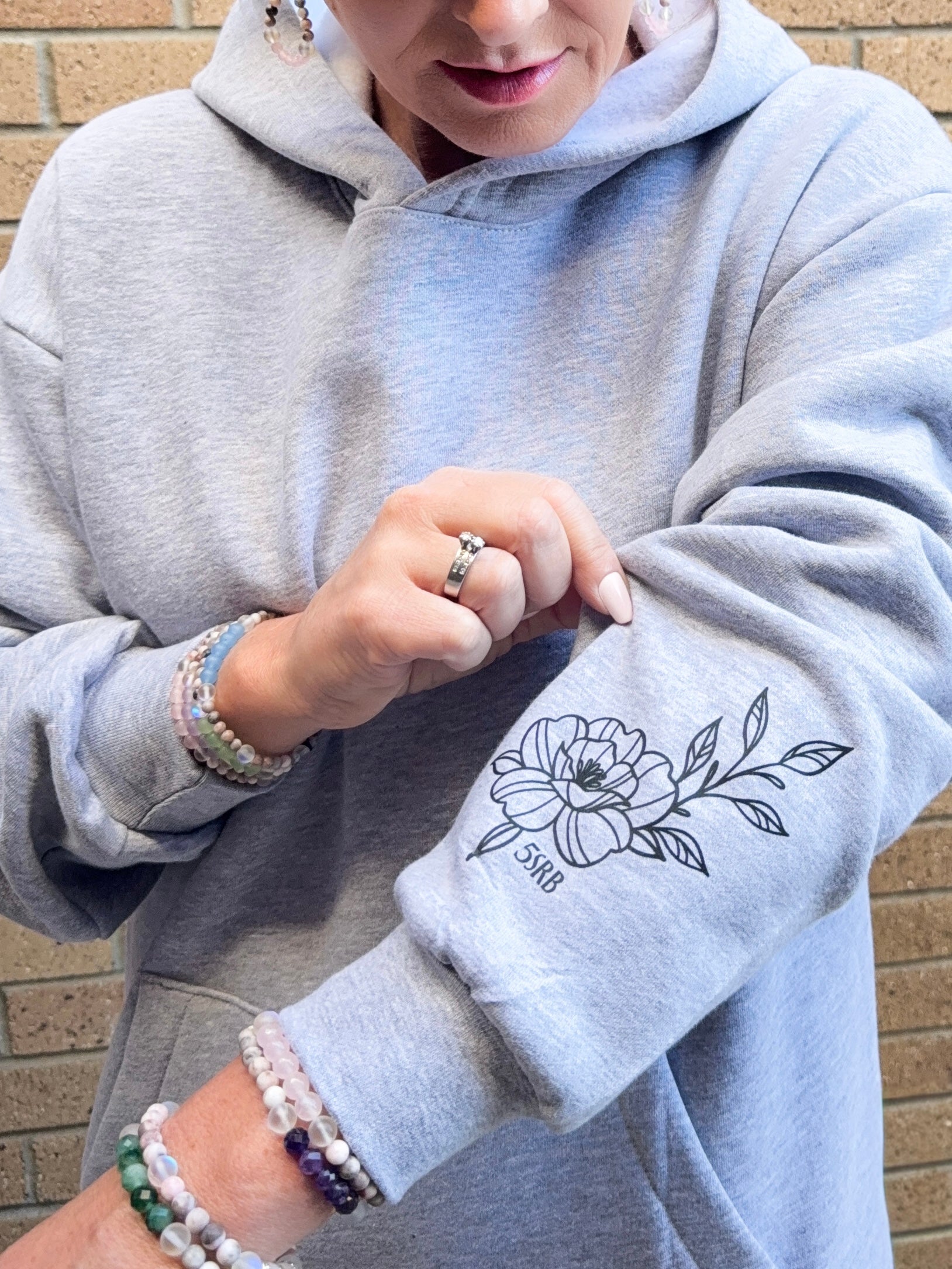 Person wearing a gray hoodie with a floral design on the sleeve, standing against a brick wall.